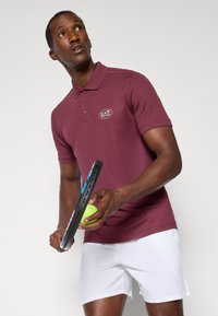 Bordo polo majica s ovratnikom i logotipom, uparena s bijelim kratkim hlačama. Osoba drži tenisku reketa i zelenu tenisku lopticu.