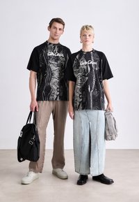 Sorte grafiske t-shirts har abstrakte mønstre og hvid branding. En model har lysebeige løse bukser på; den anden har en denimnederdel og en taske.