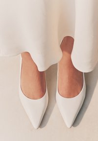 Hvide spidse flade sko med en glat lædertekstur og en lav, rundet udskæring. Skoene er placeret på en lys overflade under en kant.