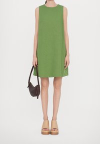 Grünes ärmelloses Kleid mit geradem Schnitt und glatter Textur. Kombiniert mit einer braunen Handtasche und beigen Plateauschuhen mit Holzsohle.