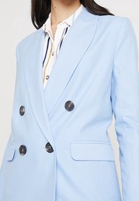 Světle modrý dvouřadý blejzr s klopami s výstřihy, černými knoflíky a dvěma předními kapsami. Nošený přes pruhované tričko. Hladká textura.