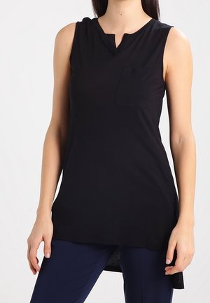 Femme portant un haut noir sans manches avec une petite poche poitrine et un pantalon bleu marine, debout les bras détendus le long du corps.