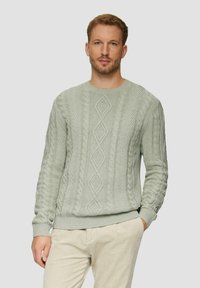 Mann mit hellbraunem Haar, der einen hellgrauen kabelgestrickten Pullover und beigetöne Hosen trägt, steht vor einem schlichten hellen Hintergrund.