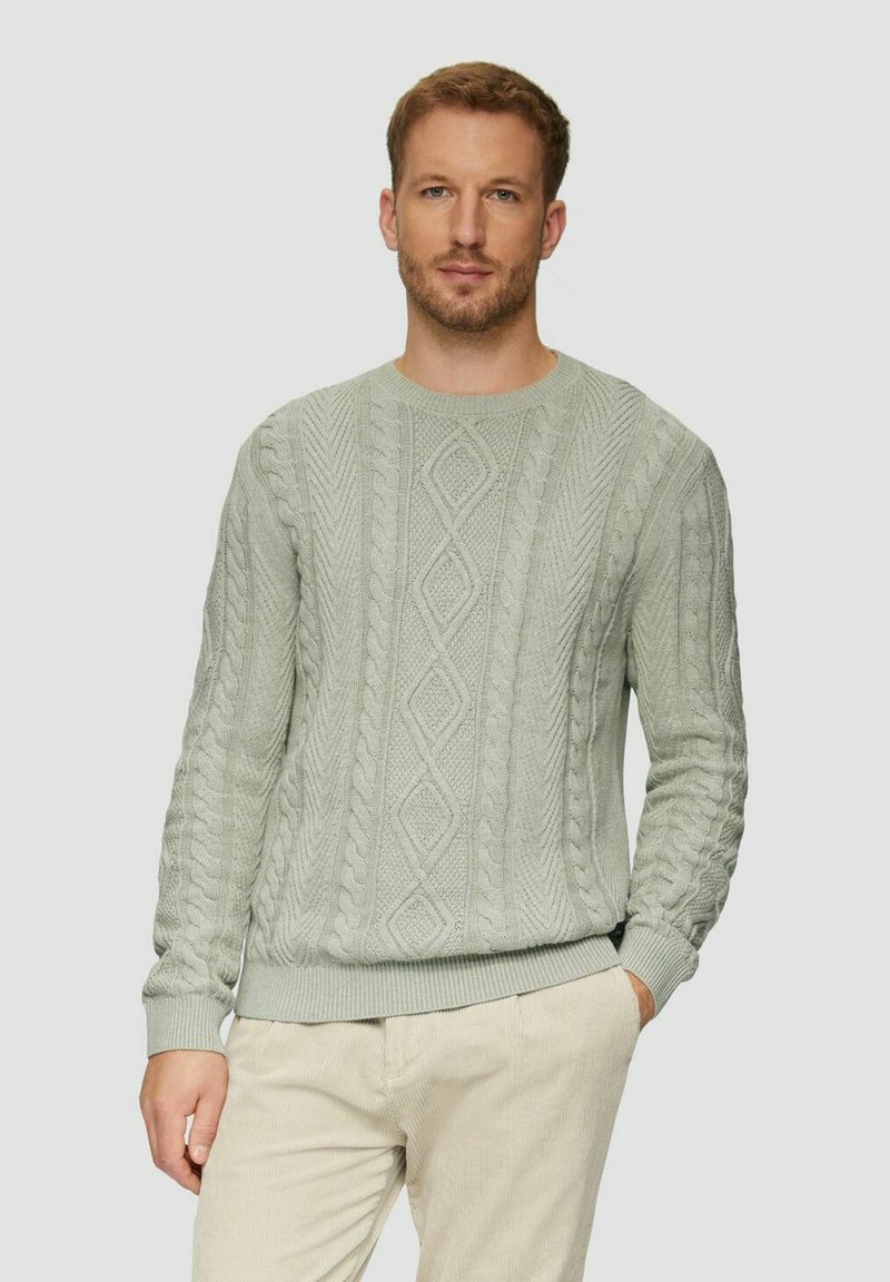 Mann mit hellbraunem Haar, der einen hellgrauen kabelgestrickten Pullover und beigetöne Hosen trägt, steht vor einem schlichten hellen Hintergrund.