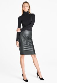 Zwart getailleerd coltruitje gecombineerd met een hooggewaste, donkergroene kokerrok van nepleer met horizontale plooien. Zwarte stilettohakken.