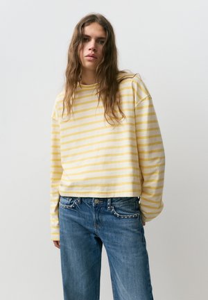Person with long wavy hair wearing a yellow and white striped long-sleeve shirt and blue jeans, standing against a plain white background.