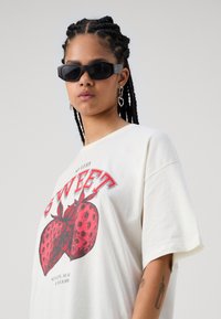 Hvid bomulds-T-shirt med et rødt jordbærmotiv og teksten "SÅ MEGET SØD". Modellen bærer sorte, kantede solbriller og sølvfarvede hjerteøreringe.