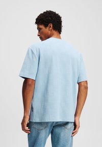 Lys blå kortærmet skjorte med struktureret stof, der har et diskret logomønster. Lige kant, rund halsudskæring, parret med falmede blå jeans.