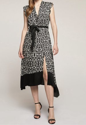 Femme portant une robe midi sans manches à motif noir et blanc, col en V, fente latérale, ceinture noire à la taille et talons noirs avec bride à la cheville.