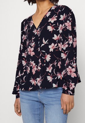 Blouse fleurie bleu marine avec des fleurs roses et blanches, encolure en V, boutons et ourlet à volants. Manches longues avec poignets élastiques.