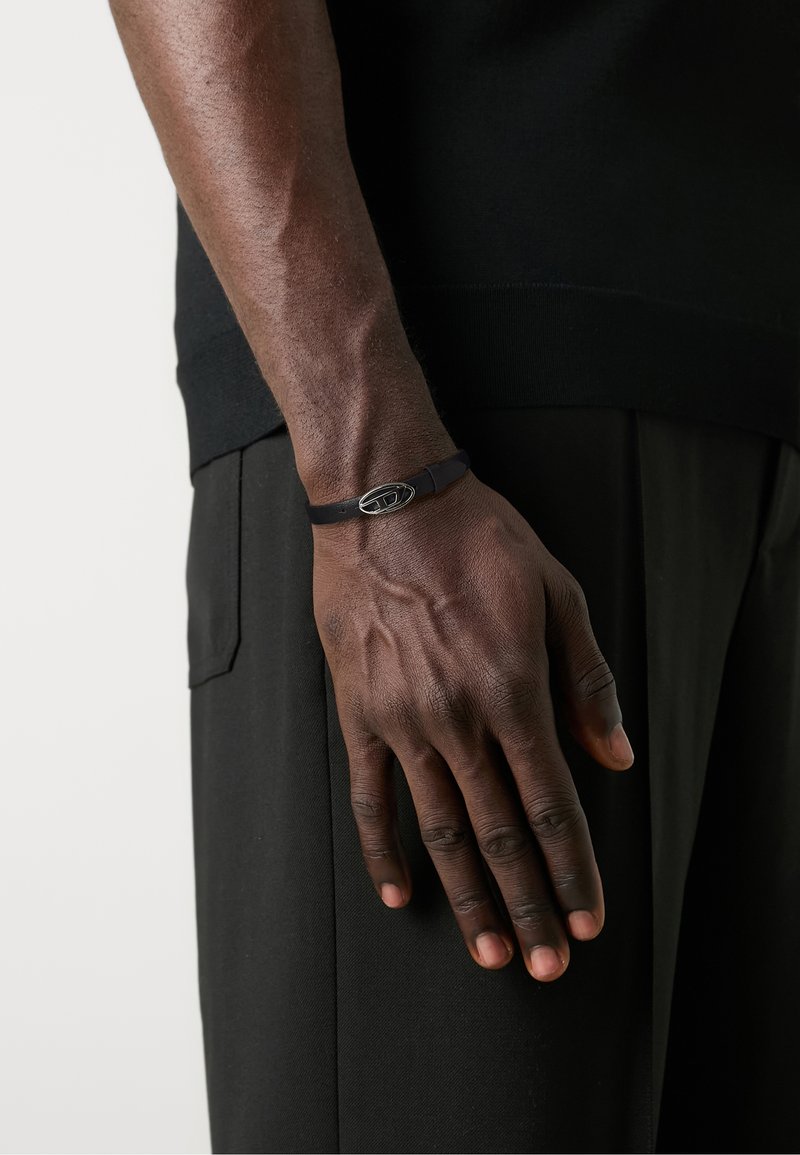 Black rubber bracelet with a silver oval clasp details, worn on a hand against dark clothing, showcasing a smooth texture and minimal design.