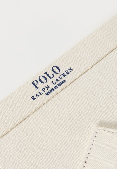Ύφασμα σε κρεμ απόχρωση με διακριτικό σχέδιο ψαροκόκκαλου, που φέρει μπλε σκούρο κείμενο "Polo Ralph Lauren Made in India" σε ραφή ρούχου.