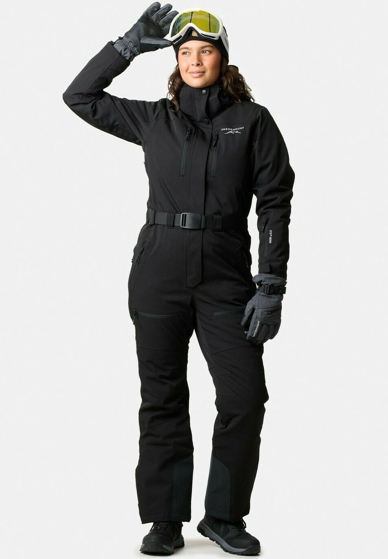 Zwarte skikleding met hoge kraag, riem, ritszakken en versterkte panelen. Vergezeld van zwarte handschoenen en goggles met gele lenzen.