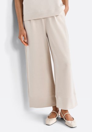 Osoba ubrana w beżowe szerokie spodnie 7/8 i pasujące beżowe płaskie buty z paskami wokół kostek, stojąca na tle białego tła.