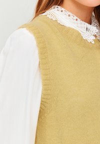Knit yellow vest with a soft texture and ribbed edges, layered over a white blouse featuring a detailed lace collar.
