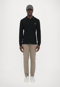 Čierna polo košeľa s dlhými rukávmi a jemne vyšitým logom, béžové slim-fit nohavice a šedá čiapka, v kombinácii s bielymi teniskami. Jednoduchý dizajn.