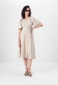 Abito beige con bottoni, maniche corte a sbuffo, vita arricciata e orlo a lunghezza ginocchio. La modella indossa sandali neri con cinturini e occhiali da sole.