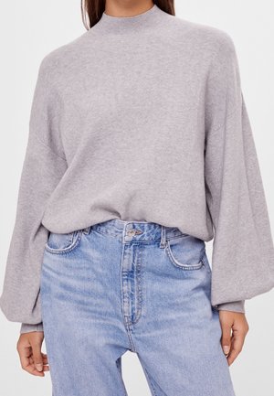 Femme portant un pull col montant à manches longues gris clair, rentré dans un jean taille haute bleu clair, les mains détendues le long du corps.