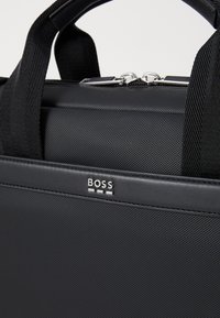 Sac noir structuré en tissu texturé avec des accents en cuir lisse. Doté de poignées supérieures, d'une fermeture éclair et d'un détail de logo sur le devant.