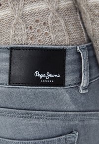 Des jeans en denim gris présentent une patte en cuir noir avec le texte blanc "Pepe Jeans LONDRES." Des détails de couture et des passants de ceinture sont visibles.