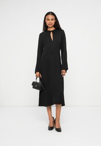 Abito midi nero con maniche lunghe e scollatura a chiave, caratterizzato da una consistenza liscia. Abbinato a una piccola borsa nera e tacchi alti.