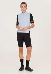 Lyseblå ærmeløs cykelvest, sorte korte ærmer, parret med sorte cykelshorts og sorte sko. Har en høj krave og tætsiddende design.