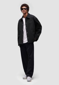 Sort oversized jakke med sølvknapper, båret over en hvid t-shirt, parret med mørke bukser og grå sneakers. Minimalistisk design, udendørsbeklædning.