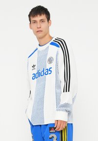 Bela pletena majica iz jerseya s plavimi in črnimi črtami, ki vključuje "adidas" blagovno znamko v modri barvi. Ima okrogel ovratnik in teksturiran vzorec.