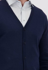 Man in een wit overhemd met een klein geometrisch patroon onder een marineblauwe gebuttonde cardigan.