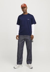 Marineblå t-skjorte med hvite langermede klær under, kombinert med mørke denimbukser og sølvfargede joggesko; en rustfarget caps fullfører antrekket.