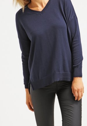 Personne portant un pull bleu marine à col en V à manches longues et un pantalon noir ajusté, debout sur un fond blanc.