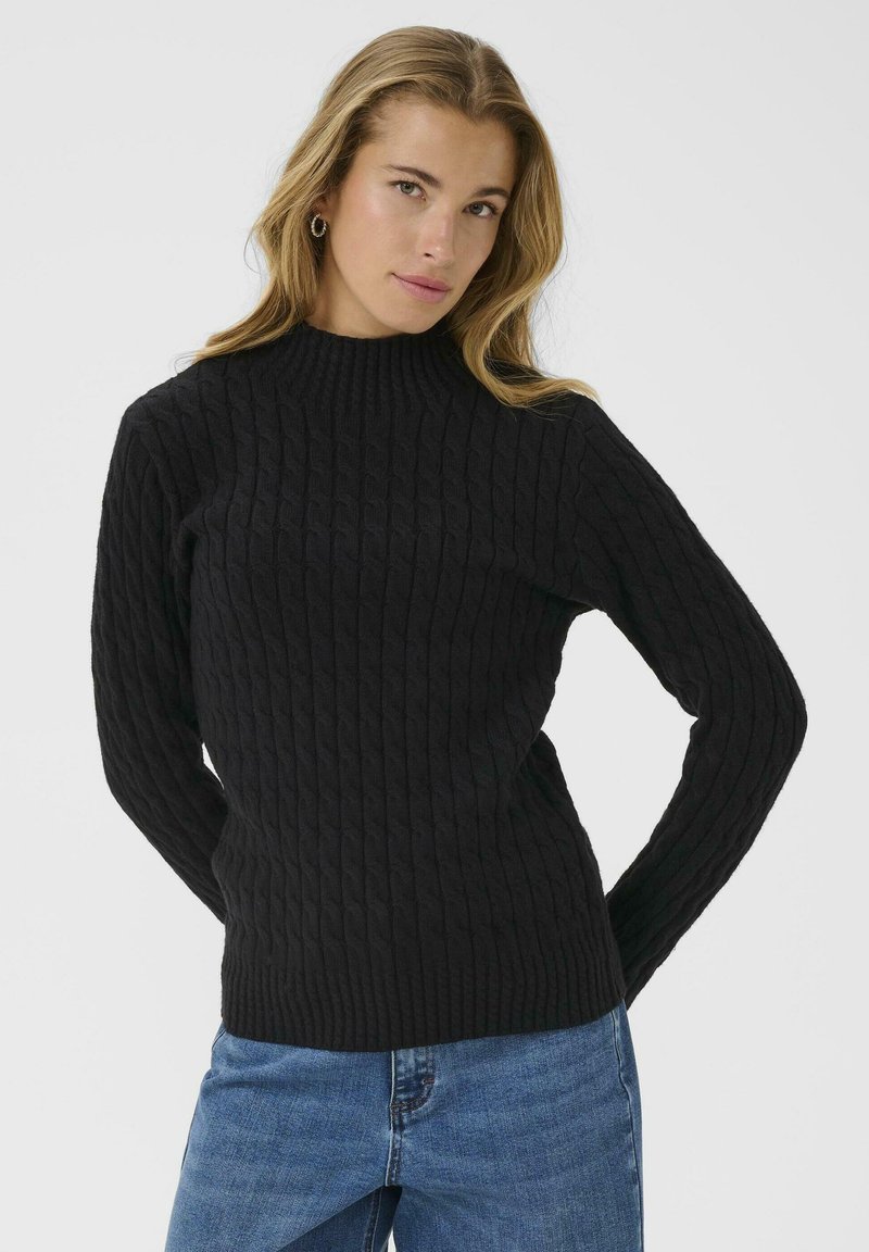 Zwarte geribbelde gebreide trui met hoge hals en lange mouwen, met een textuurpatroon. Gecombineerd met blauwe denim jeans.
