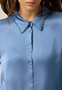 Camisa azul claro con cuello clásico, que presenta un ribete decorativo y cierres de botón. Textura de tejido suave, ligero brillo y dos botones visibles.