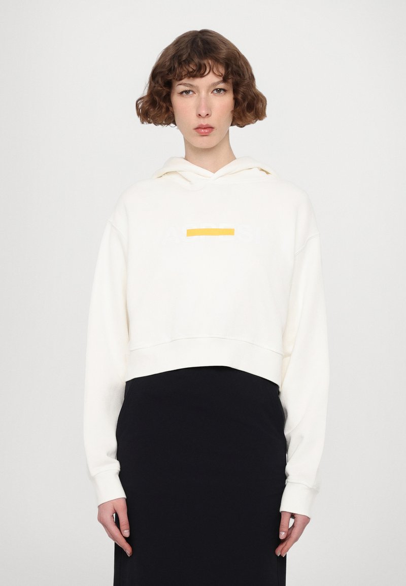 Haut à capuche court blanc avec un graphique avant en gris clair et un petit accent jaune. Il comporte un cordon de serrage à la capuche et des manches longues.