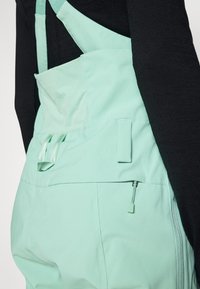 Los overoles color verde menta cuentan con tirantes ajustables, un bolsillo frontal y una cremallera lateral. La tela parece suave con un acabado mate.