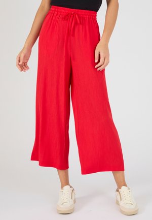 Mujer con pantalones cortos de pierna ancha de color rojo brillante con cintura ajustable y zapatillas blancas con plataforma, de pie frente a un fondo liso.