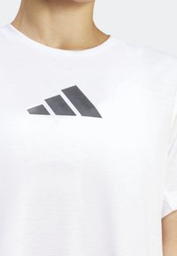 Camiseta blanca con un logo de tres rayas negro impreso en el pecho. El material parece suave; el diseño es sencillo con un cuello redondo.