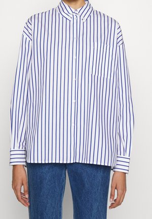 Person wearing a white long-sleeve button-up shirt with vertical blue stripes and blue jeans, standing against a plain background.