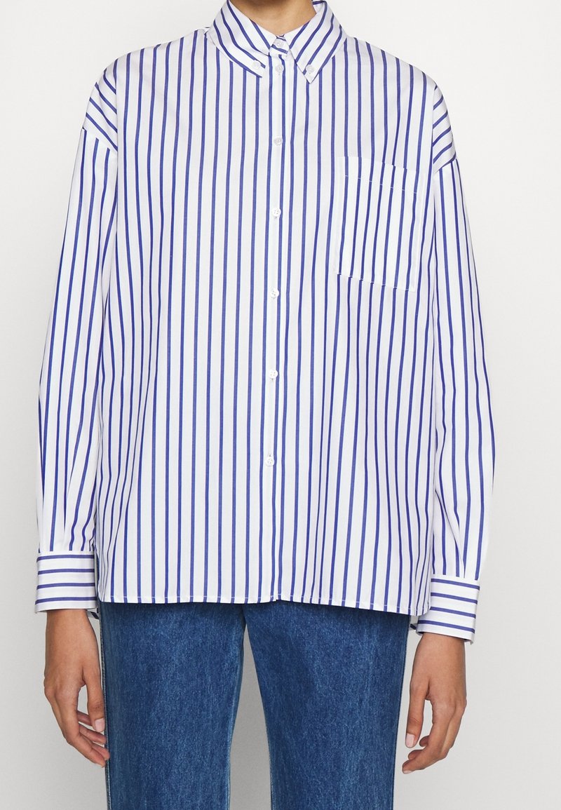 Personne portant une chemise blanche à manches longues avec des rayures verticales bleues et un jean bleu, debout devant un fond uni.