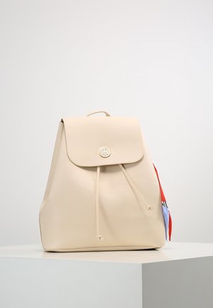 Beige leren rugzak met klep en trekkoordsluiting, gouden embleem en een kleurrijk stoffen lint aan de zijkant, getoond op een witte ondergrond.