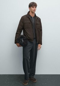 Young man wearing a dark brown jacket, black pants, and brown shoes, holding a rolled-up dark bag against a light gray wall.