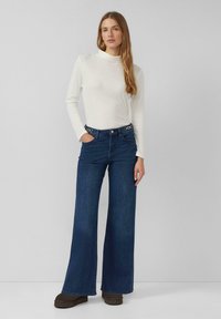 Witte top met lange mouwen gecombineerd met donkerblauwe jeans met wijde pijpen, zilverkleurige hardware accenten en zijzakken. Model staat met handen in de zakken.