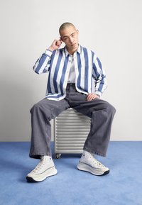 Randig blå och vit skjorta med knappar, grå säckiga byxor och vita hög-top sneakers, sittande på en silver metallisk låda mot en ljus bakgrund.