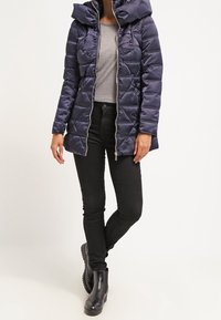 Marineblå dunjakke med hætte, lynlås lukning, quiltet design og sidelommer. Bæres over en grå skjorte og kombineres med sorte bukser og støvler.