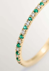 Anillo de oro con engastes alternos de piedras preciosas verdes y transparentes a lo largo de la banda, mostrando una ligera curvatura y un acabado pulido.