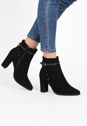 Bottines en daim noir avec un bout pointu, un large talon, des clous décoratifs et un détail de zip sur le côté. Accentuées par une sangle et une boucle.