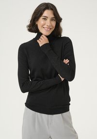 Pull noir à col roulé en tissu tricoté scintillant, avec une coupe ajustée et des poignets côtelés, porté avec un pantalon gris clair.