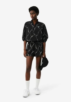Femme debout portant un t-shirt à manches longues noir à motif marbré et un short assorti, chaussettes blanches, baskets blanches, tenant une casquette noire dans une main.