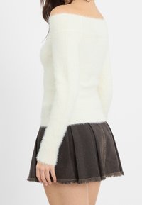 Donna che indossa un maglione bianco a spalla scoperta e una gonna nera corta a pieghe con orlo sfrangiato, mostrata di lato e di retro.