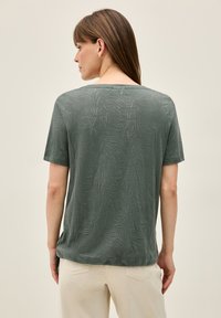 Top verde de manga corta con un patrón texturizado de hojas, escote redondo y corte holgado, combinado con pantalones de color claro. Se muestra la vista posterior.